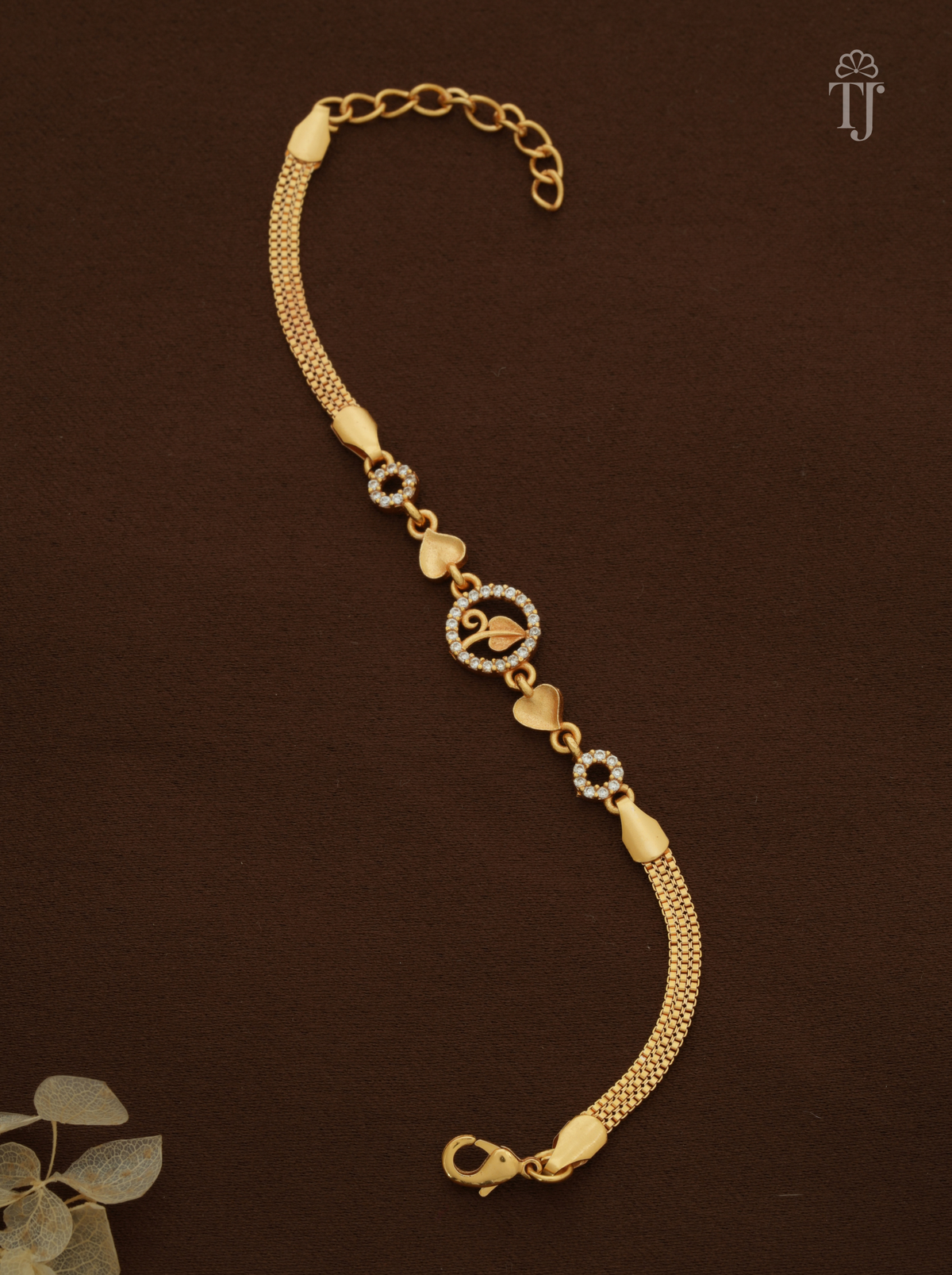 A gold-plated chain bracelet with heart and flower motifs, displayed on a dark brown background with a small decorative leaf in the corner.
