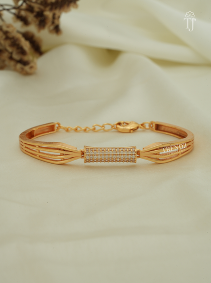 A gold-plated bracelet with a central pavé-set rectangular bezel and geometric side links, displayed on draped cream fabric with dried flowers in the background.