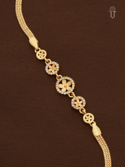 A gold chain bracelet featuring five circular floral motifs, with three adorned with small clear stones, displayed diagonally on a dark brown textured background.