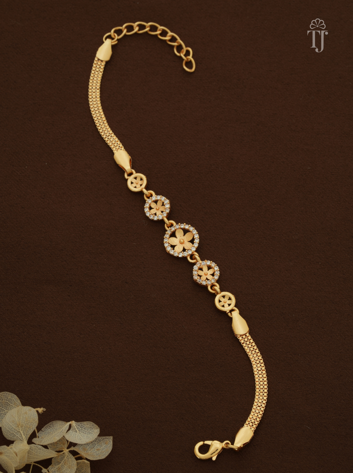 A gold-plated chain bracelet with decorative floral and geometric motifs, displayed on a dark brown background with a small decorative leaf in the corner.