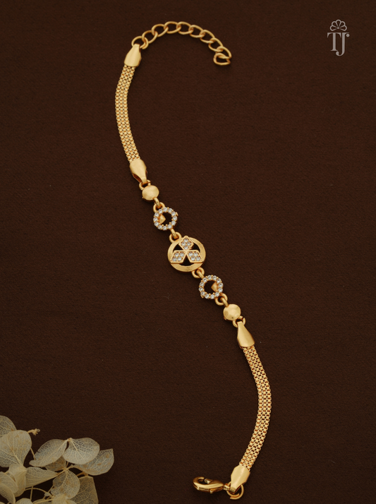 A gold-plated chain bracelet with a central circular motif and two circular links set with small clear stones, displayed on a dark brown background with a decorative eucalyptus leaf in the corner.