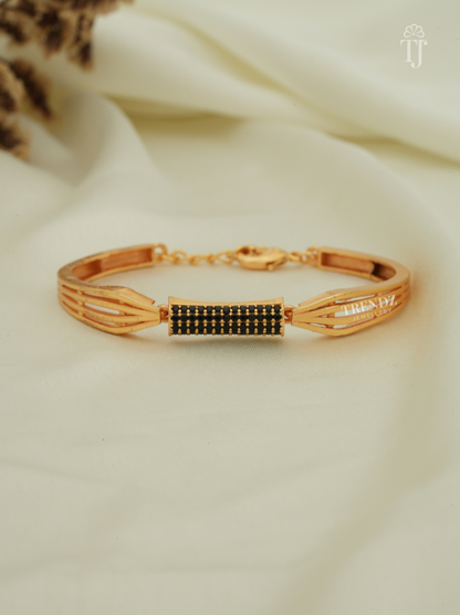 A gold-plated bangle bracelet with a central rectangular section covered in small black stones, displayed on a soft cream-colored fabric background.
