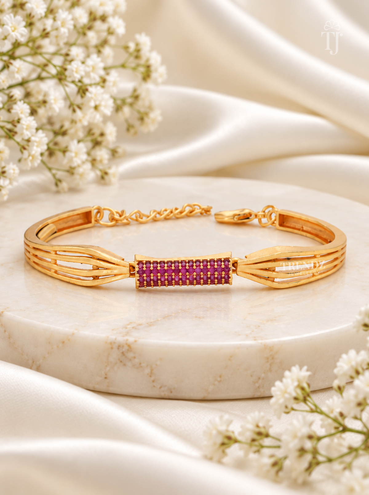 A gold-plated multi-strand bracelet with a central rectangular inlay of small pinkish-purple stones, displayed on a round white marble slab with white satin fabric and baby's breath flowers.