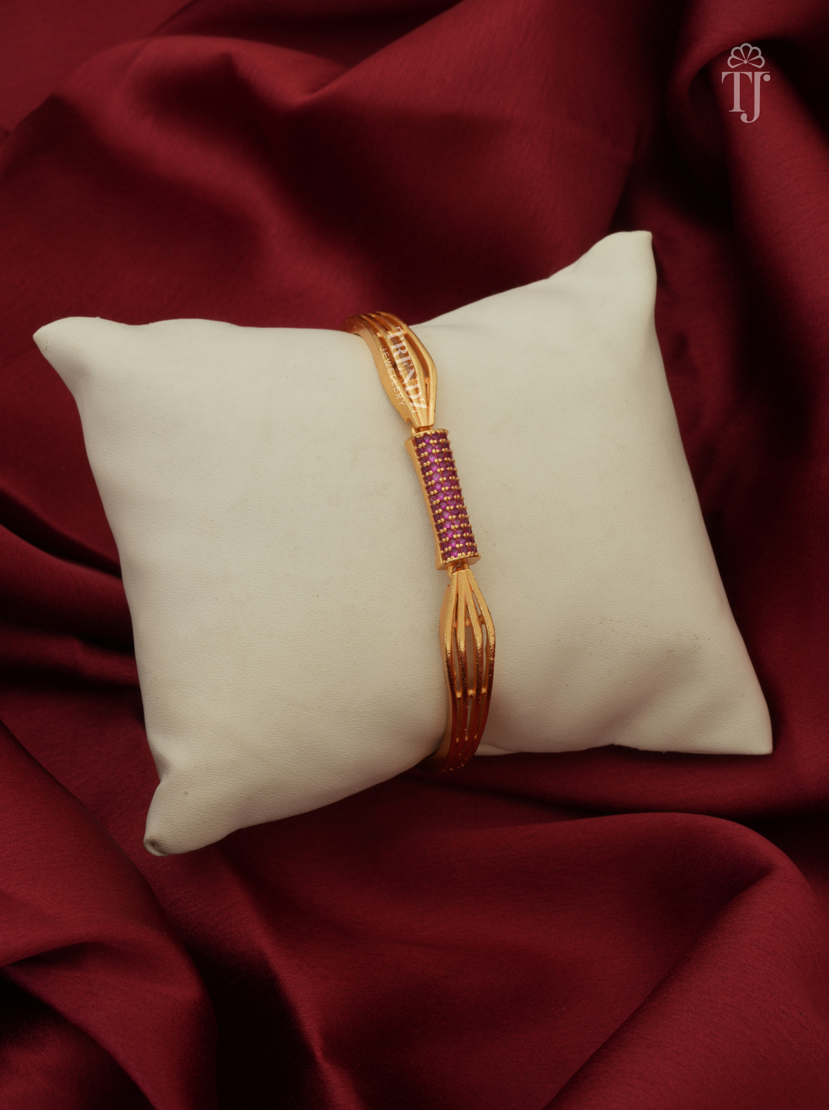 A gold-plated bangle bracelet with a central pinkish-red stone-encrusted section, displayed on a cream pillow over a draped dark red fabric background.