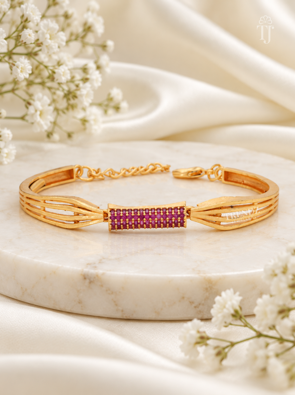A gold-plated bangle bracelet with a central rectangular bezel-set row of deep pinkish-purple stones, displayed on a white marble slab with white satin fabric and baby's breath flowers.