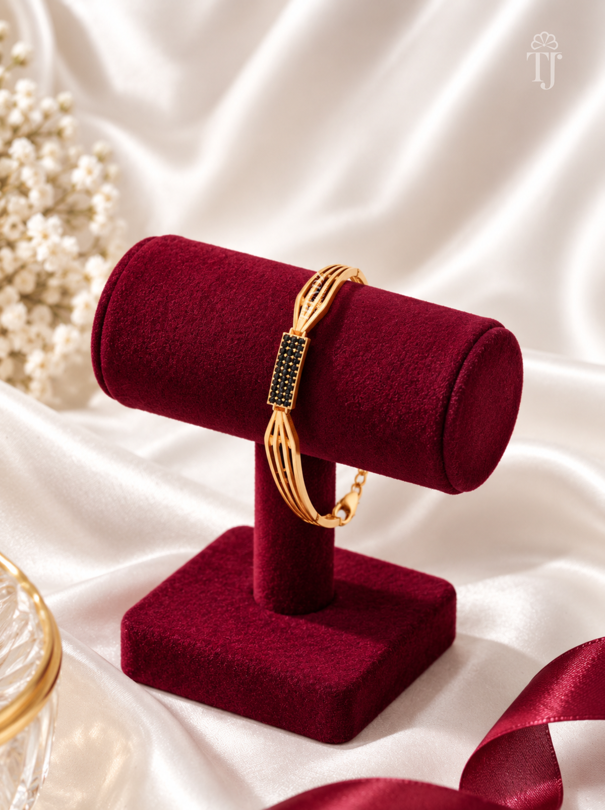 A gold-toned bangle bracelet with a rectangular black stone-set centerpiece, displayed on a deep burgundy velvet jewelry stand against a white satin backdrop.