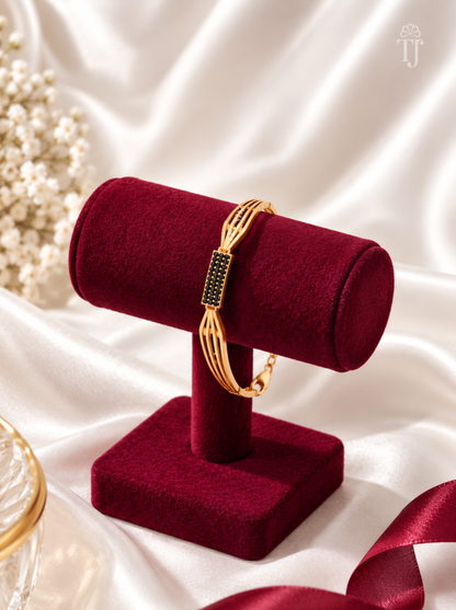 A gold-toned bangle bracelet with a rectangular black stone-set centerpiece, displayed on a deep burgundy velvet jewelry stand against a white satin backdrop.