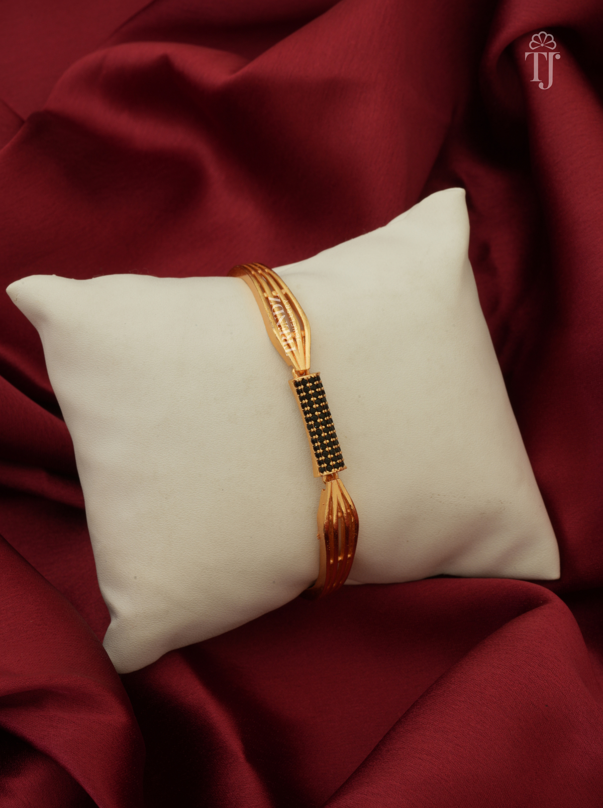 A gold-plated bangle bracelet with a black beaded center section, displayed on a cream-colored square pillow over a draped dark red satin fabric background.