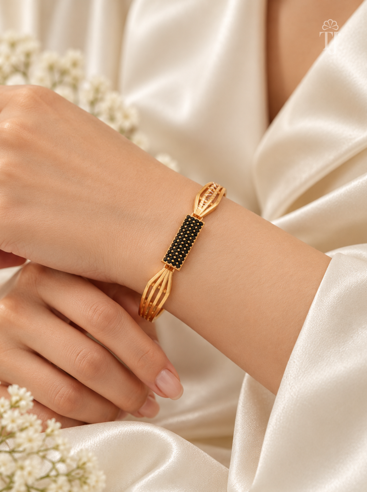 A gold-toned bangle bracelet with a central black beaded rectangular section, worn on a person's wrist. The person is wearing a cream satin robe, with small white flowers in the foreground.