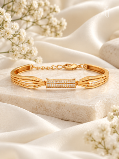 A gold-toned bracelet with a central pavé-set rectangular bar, displayed on a light stone slab with draped white fabric and small white flowers around it.