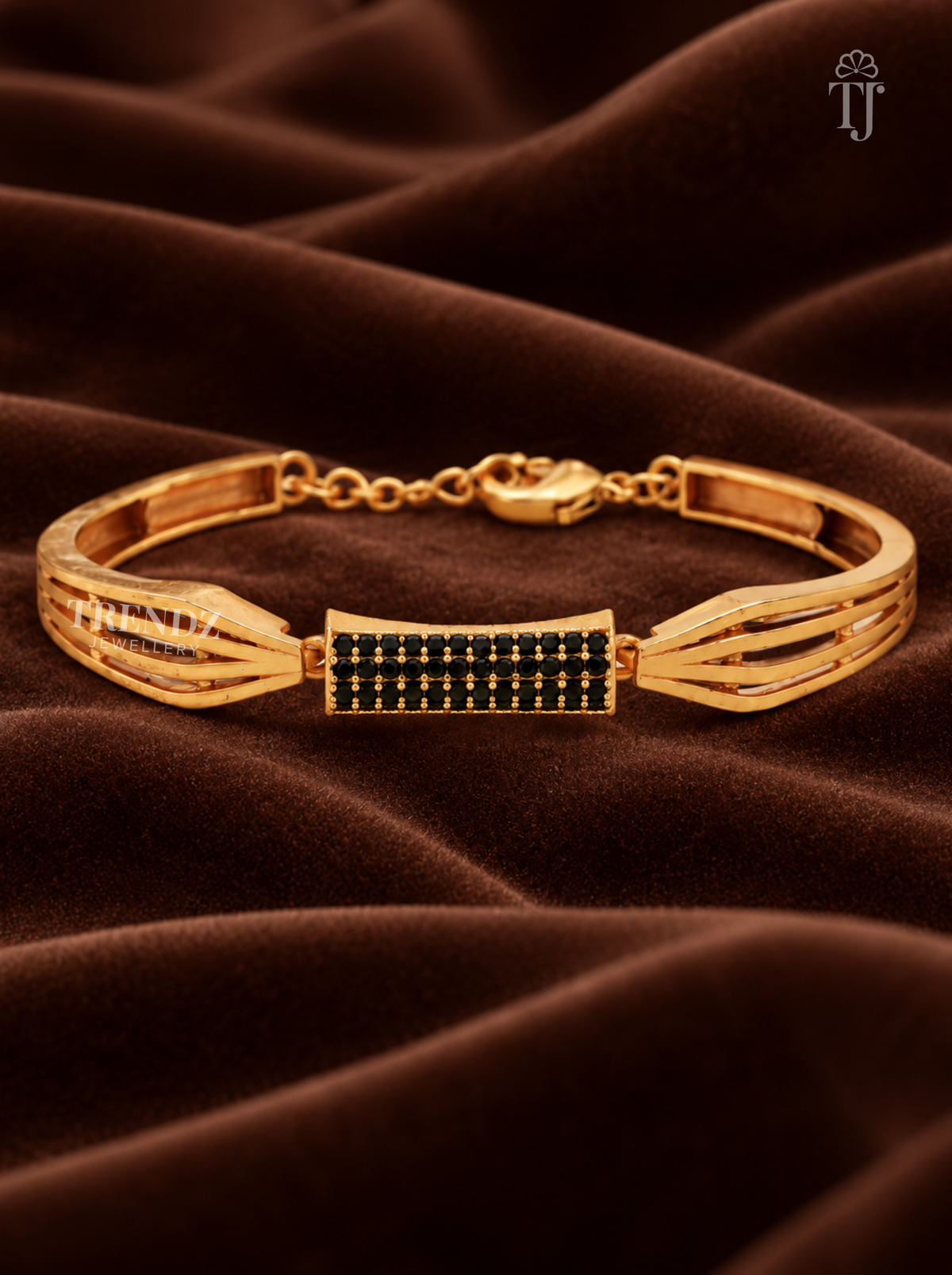 A gold-plated bangle bracelet with a central rectangular panel set with small black stones, displayed on a draped brown velvet fabric background.