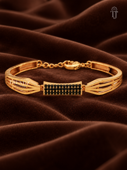 A gold-plated bangle bracelet with a central rectangular panel set with small black stones, displayed on a draped brown velvet fabric background.