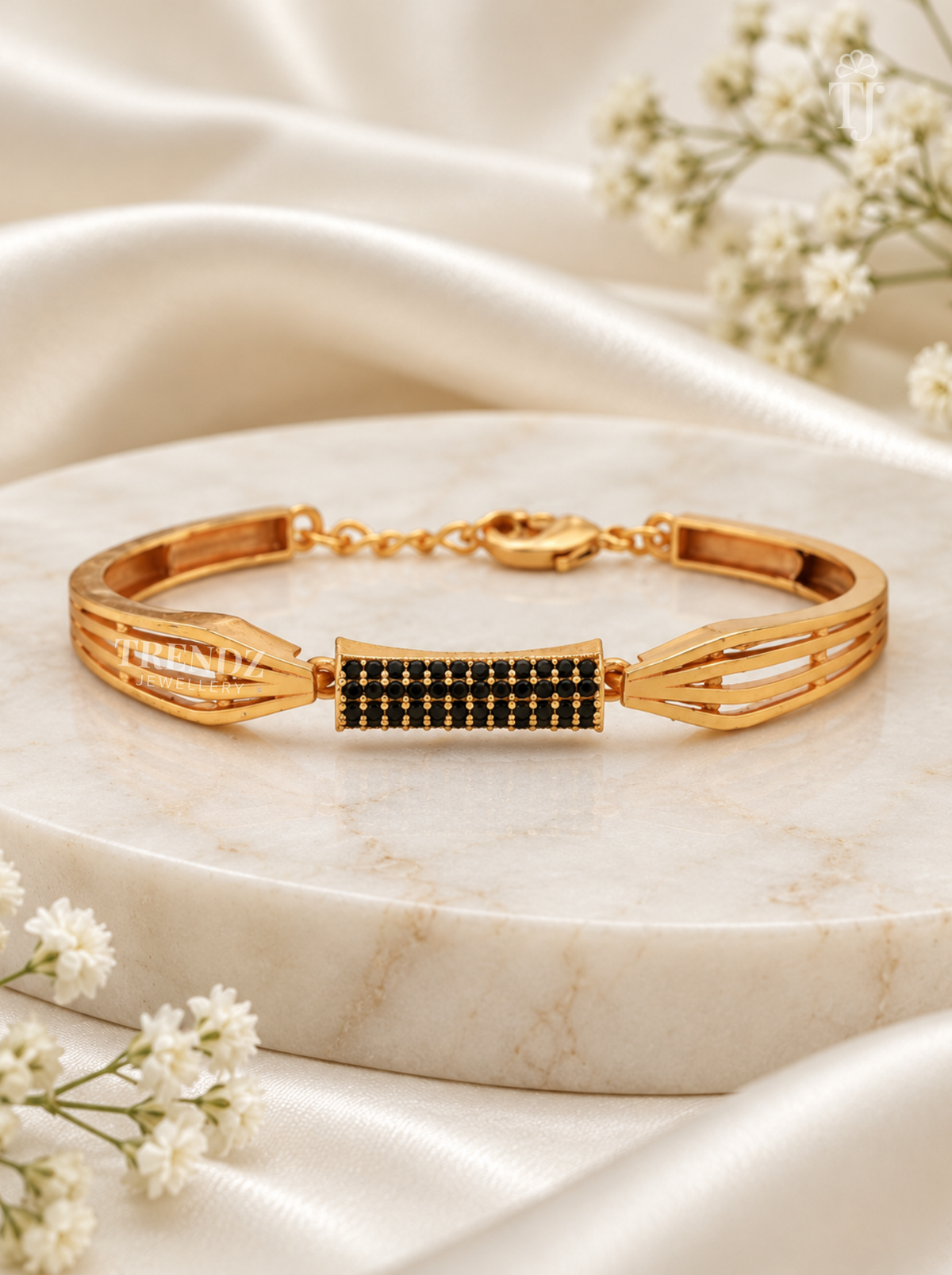 A gold-toned bangle bracelet with a central rectangular black stone inlay, displayed on a round white marble slab with white satin fabric and small white flowers around it.