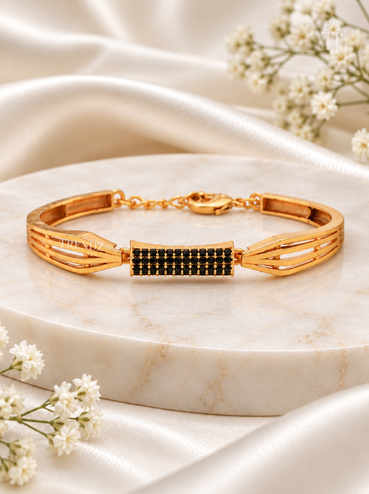 A gold-toned bracelet with a central rectangular section set with small black stones, displayed on a round white marble slab with draped satin fabric and small white flowers around it.
