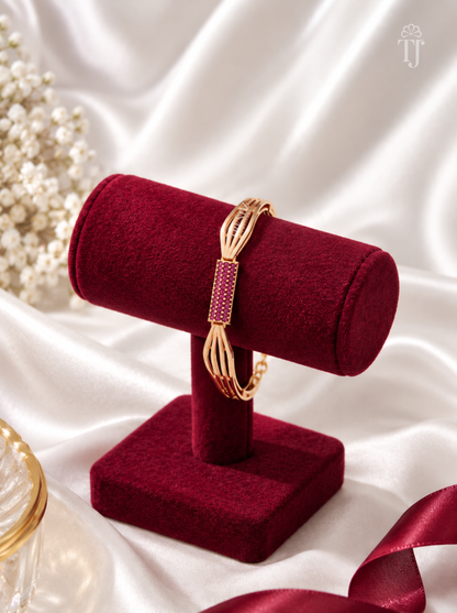 A gold-toned bangle bracelet with a rectangular pinkish-red stone-set center, displayed on a deep red velvet jewelry stand against a white satin backdrop.