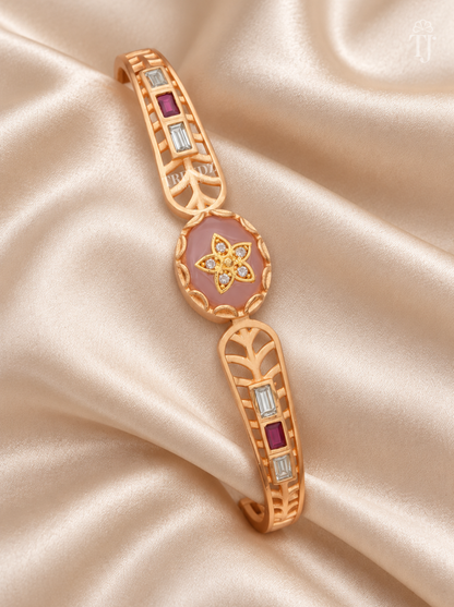 A gold-toned cuff bracelet with a pink enamel center, a floral motif, and rectangular red and clear stones, displayed on draped champagne satin fabric.