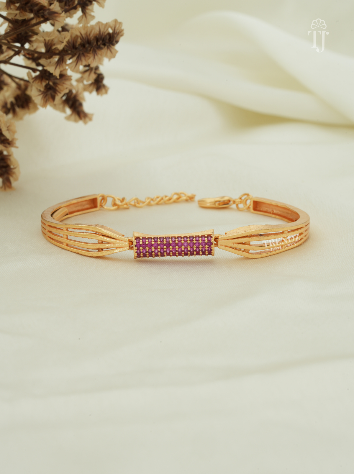 A gold-plated bangle bracelet with a central rectangular section paved with small pinkish-purple stones, displayed on a soft cream fabric background with dried flowers in the corner.