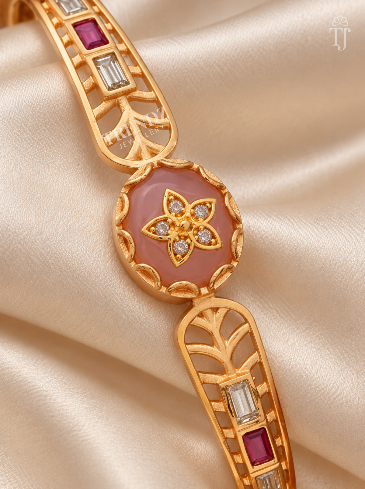 A gold-tone bracelet with a central oval pink stone surrounded by small clear stones and a floral motif, displayed on draped champagne satin fabric.