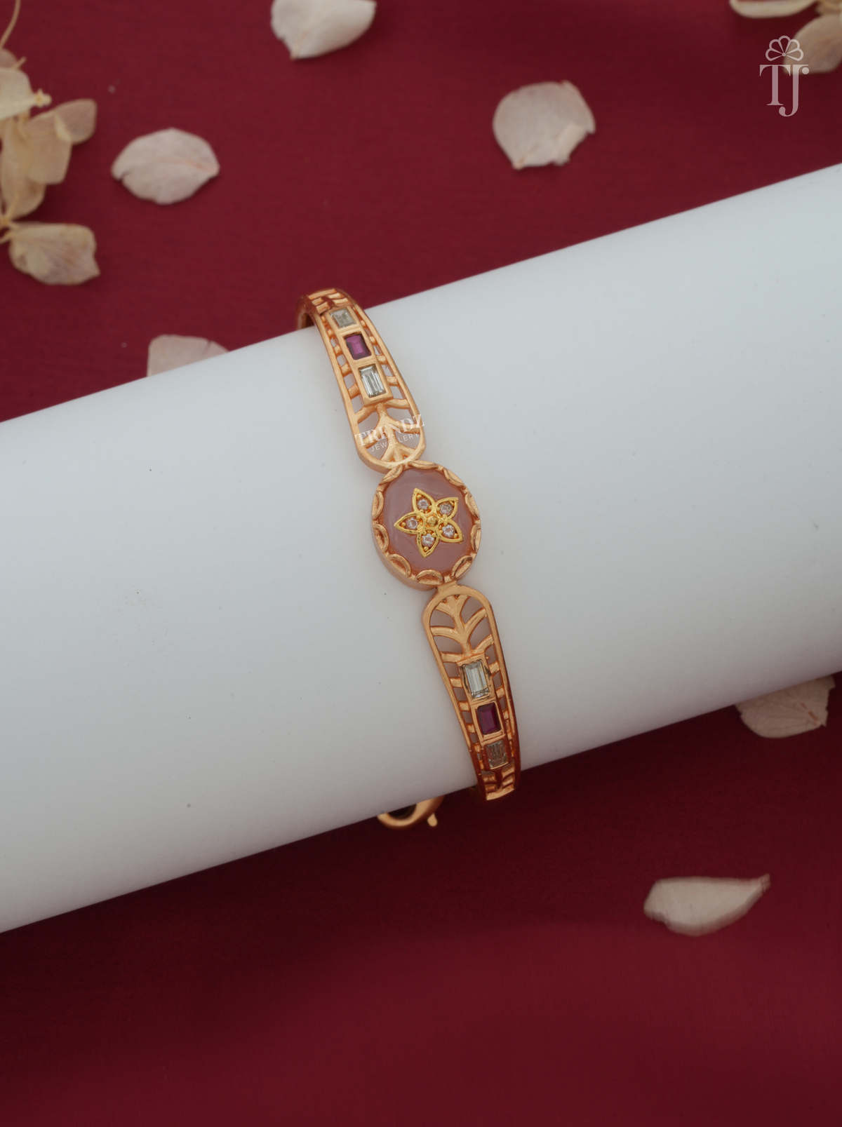 A gold-plated traditional Indian bangle bracelet with red enamel and small colored stone accents, displayed on a white cylindrical stand against a dark red background with scattered dried flower petals.