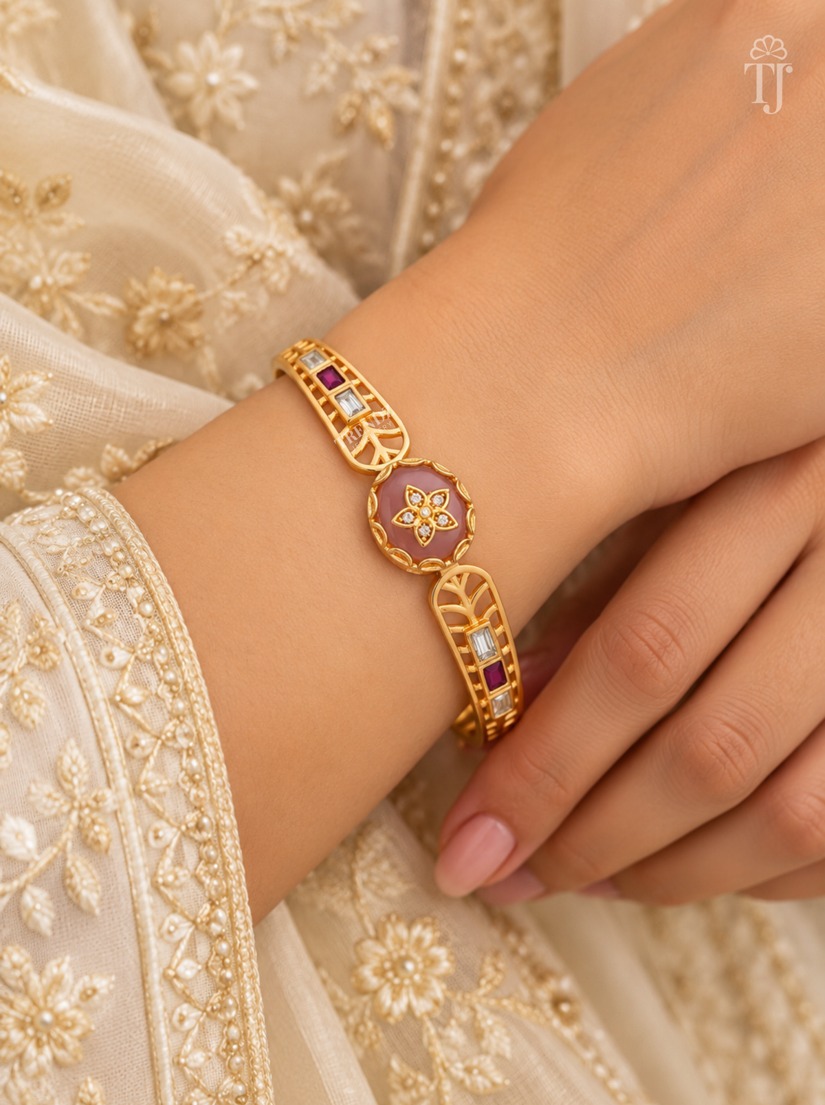 A gold-toned traditional bracelet with a central oval purple enamel panel and small red and clear stones, shown worn on a person's wrist against an embroidered cream garment.