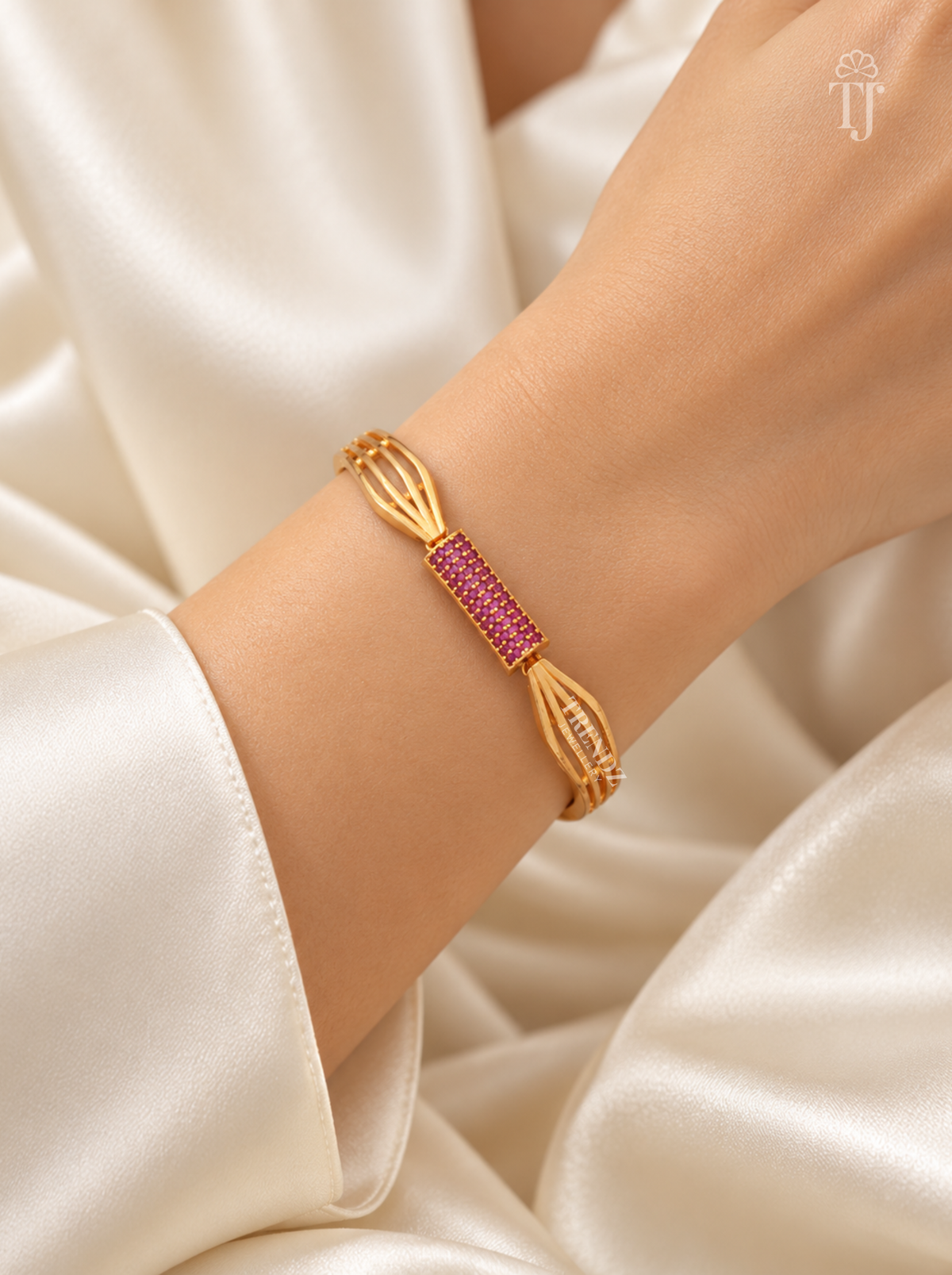 A gold-toned bangle bracelet with a central rectangular section paved with pink stones, shown worn on a person's wrist against a soft cream satin fabric background.