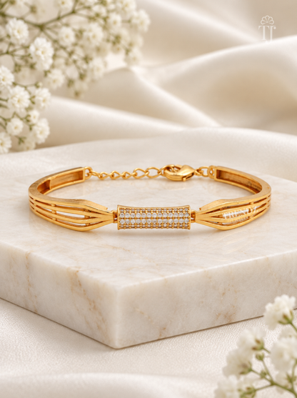 A gold-toned bracelet with a central pavé rectangular section and matching pavé details on the side, displayed on a white marble block with soft white fabric and small white flowers in the background.
