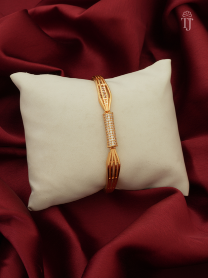 A gold-toned bangle bracelet with a central pavé-set stone section, displayed on a cream pillow over draped dark red satin fabric. A small 'TJ' logo watermark appears in the top right corner.