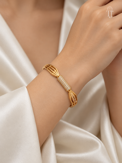 A gold-toned bangle bracelet with a central row of small white stones, worn on a model's wrist. The model is wearing a cream satin robe, and a small 'TT' logo is visible in the top right corner.