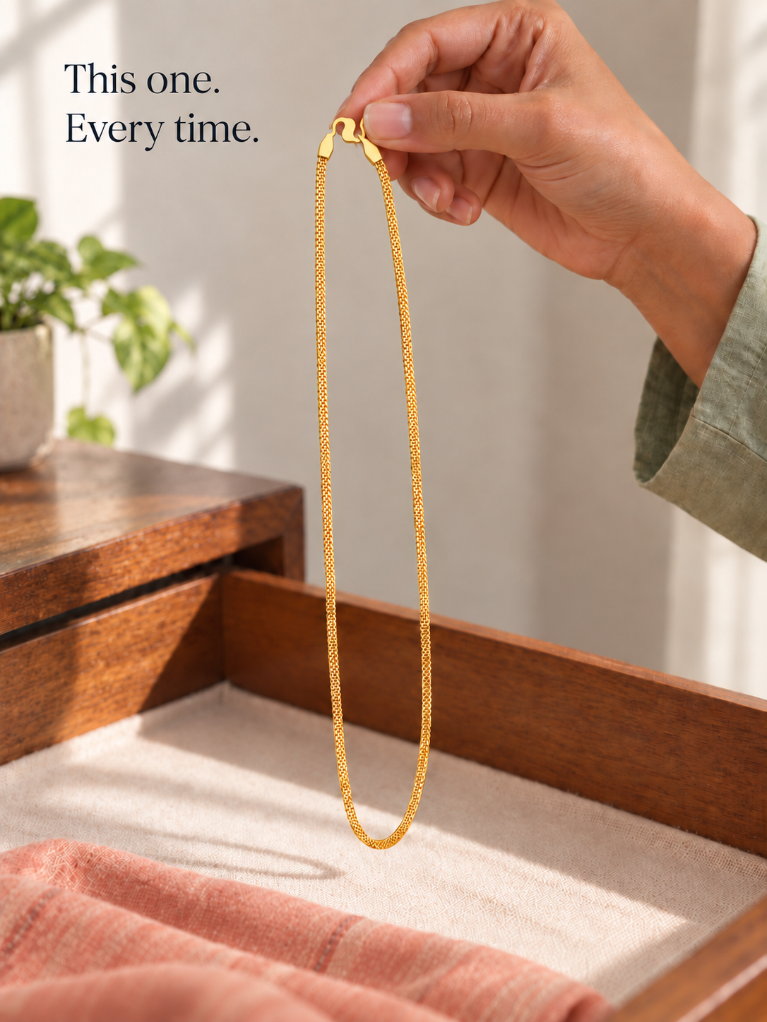 Hand holding thin gold plated box link chain above wooden jewellery tray