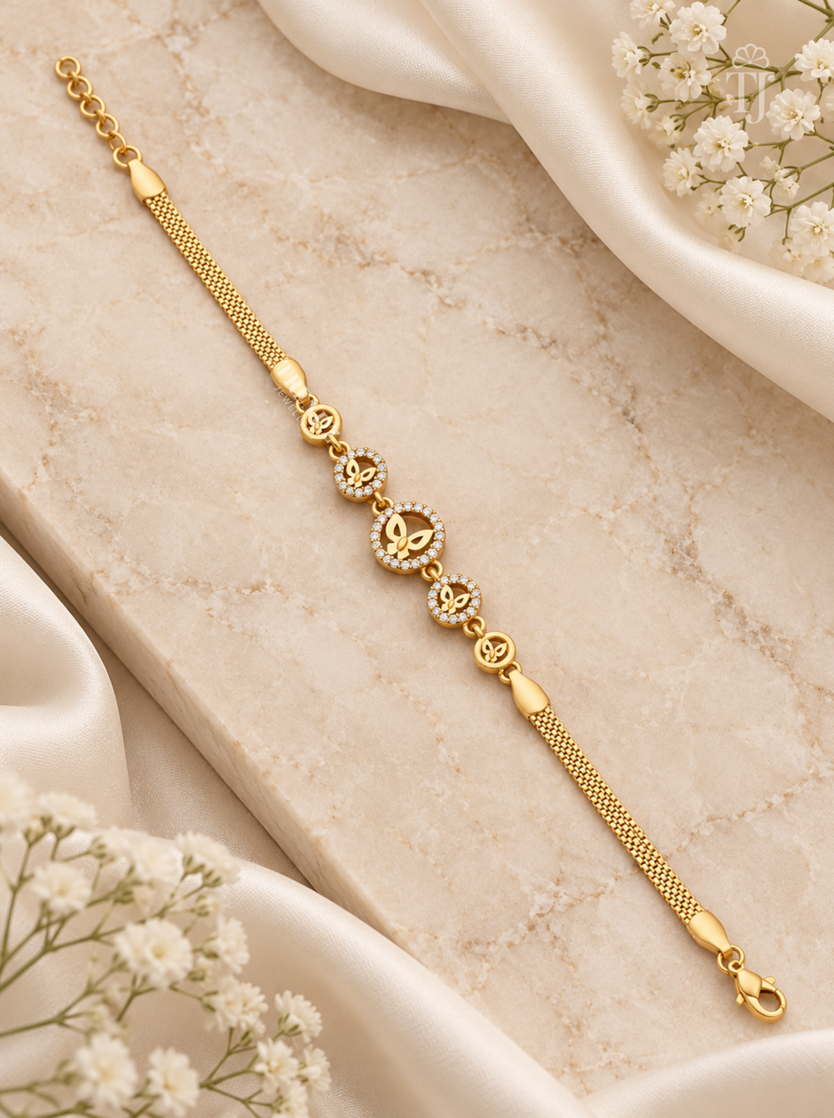 A gold chain bracelet with a central heart-shaped cluster of small clear stones and repeating heart motifs, styled on a beige marble slab with white satin and baby's breath flowers.