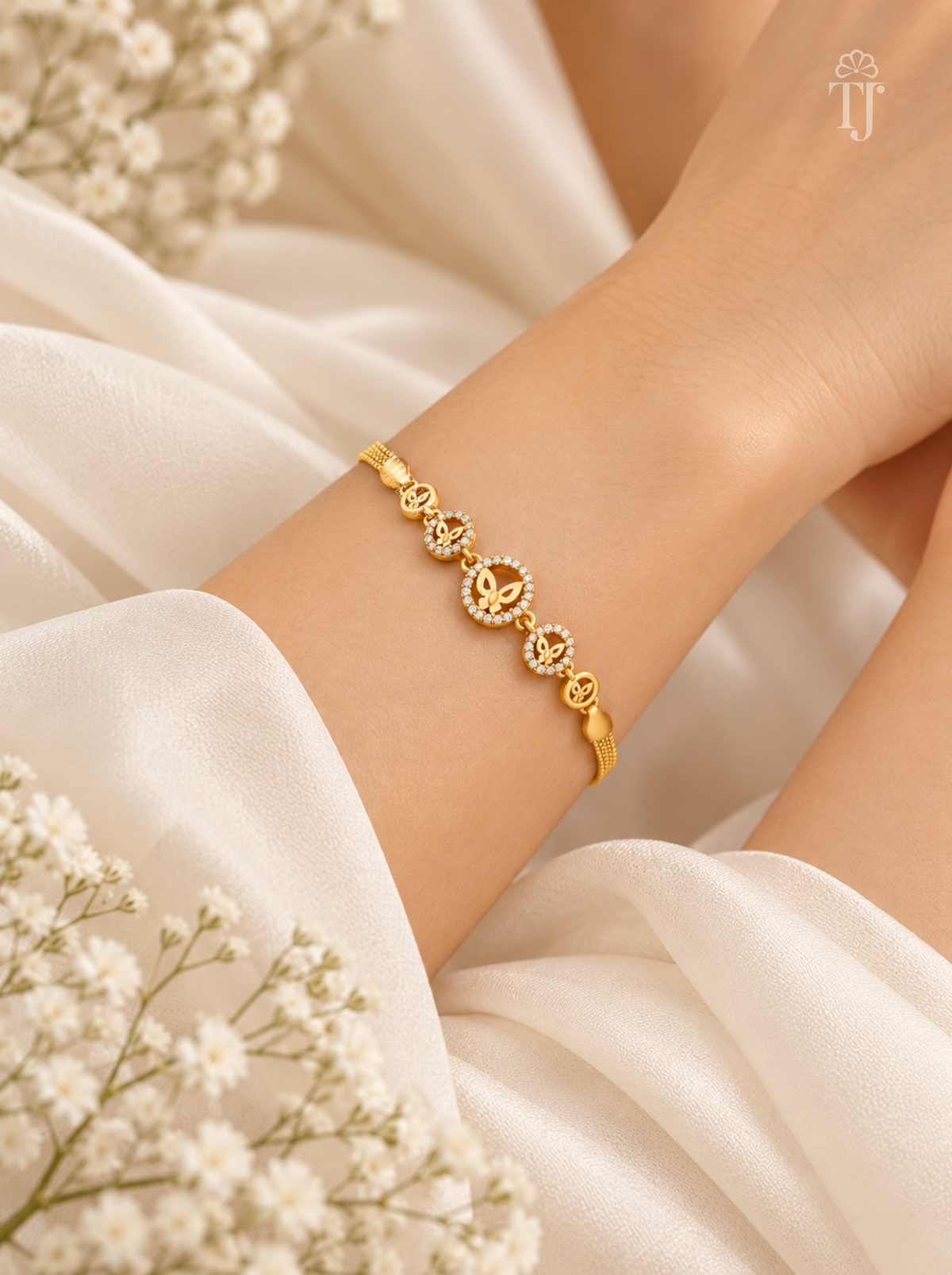 A delicate gold bracelet with circular floral motifs and small clear stones, worn on a model's wrist against a soft white fabric background with baby's breath flowers.