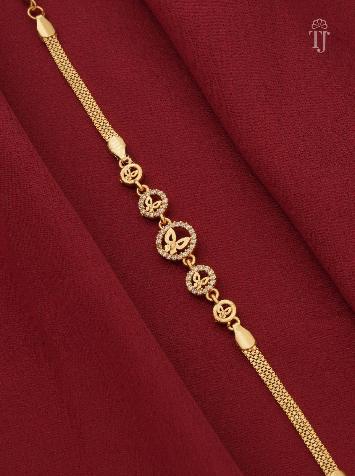A gold chain bracelet with circular butterfly and monogram motifs, displayed diagonally on a draped dark red fabric background.