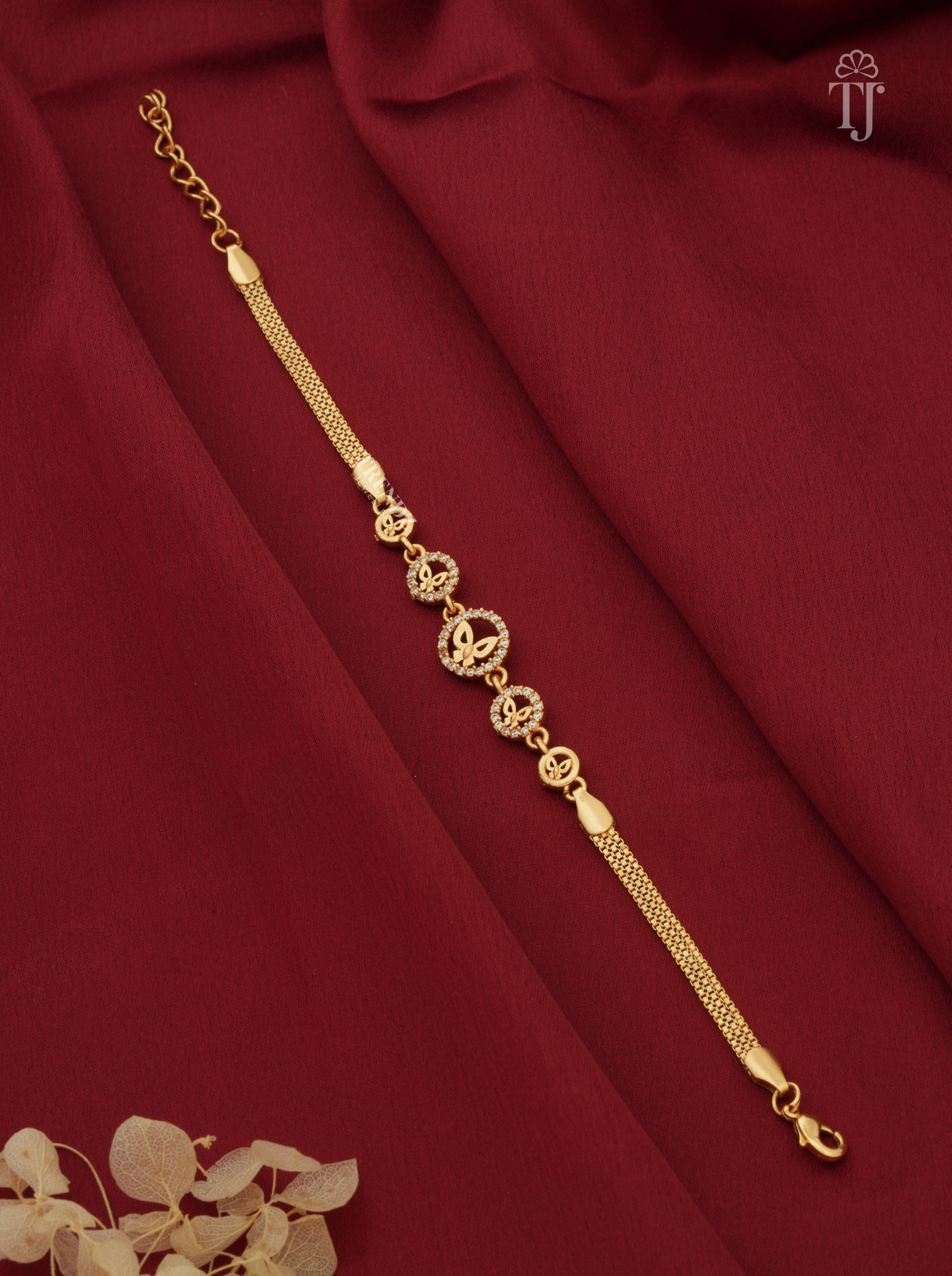 A gold-toned chain anklet with ornate circular and floral motifs, laid diagonally on draped dark red fabric with a small dried floral accent in the corner.