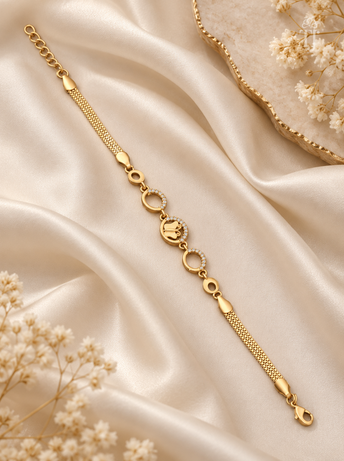A delicate gold chain bracelet with a central oval charm, displayed on draped champagne satin fabric with dried baby's breath flowers.
