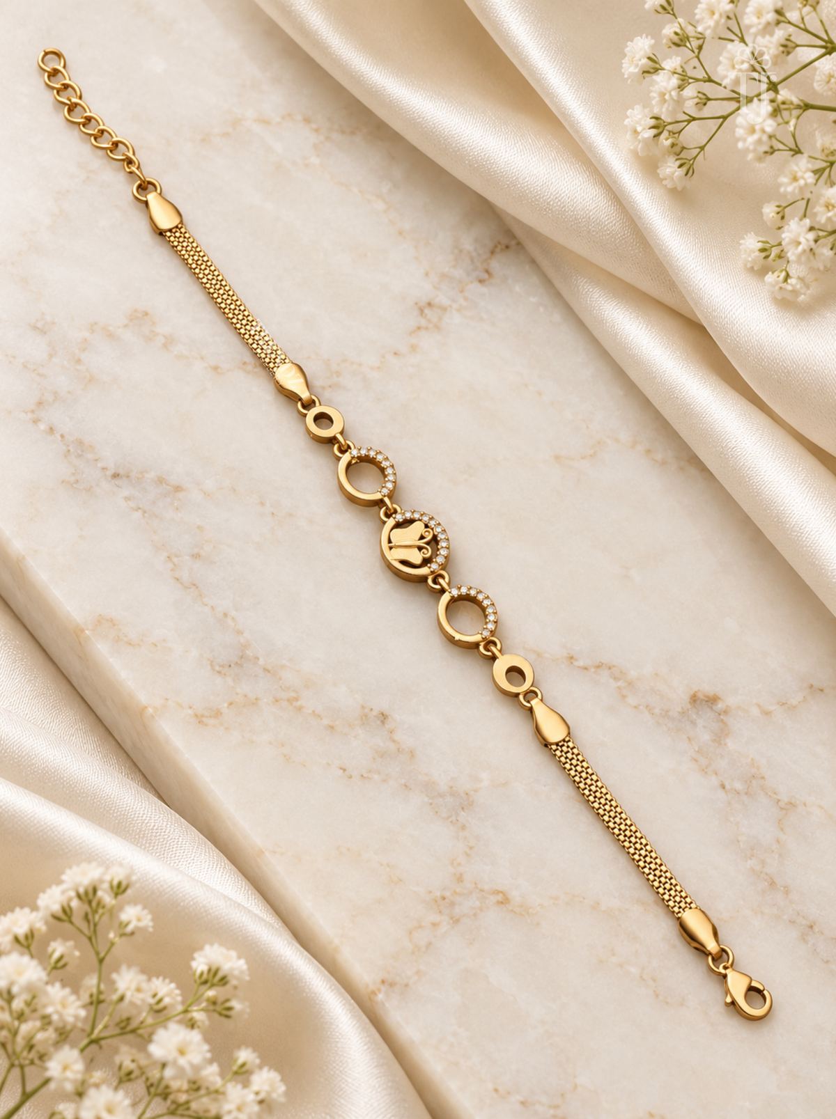 A gold chain bracelet with a central oval charm featuring a crown motif and small circular details, styled on a cream marble surface with draped satin and white baby's breath flowers.