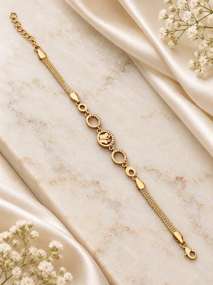 A gold chain bracelet with a central oval charm featuring a crown motif and small circular details, styled on a cream marble surface with draped satin and white baby's breath flowers.