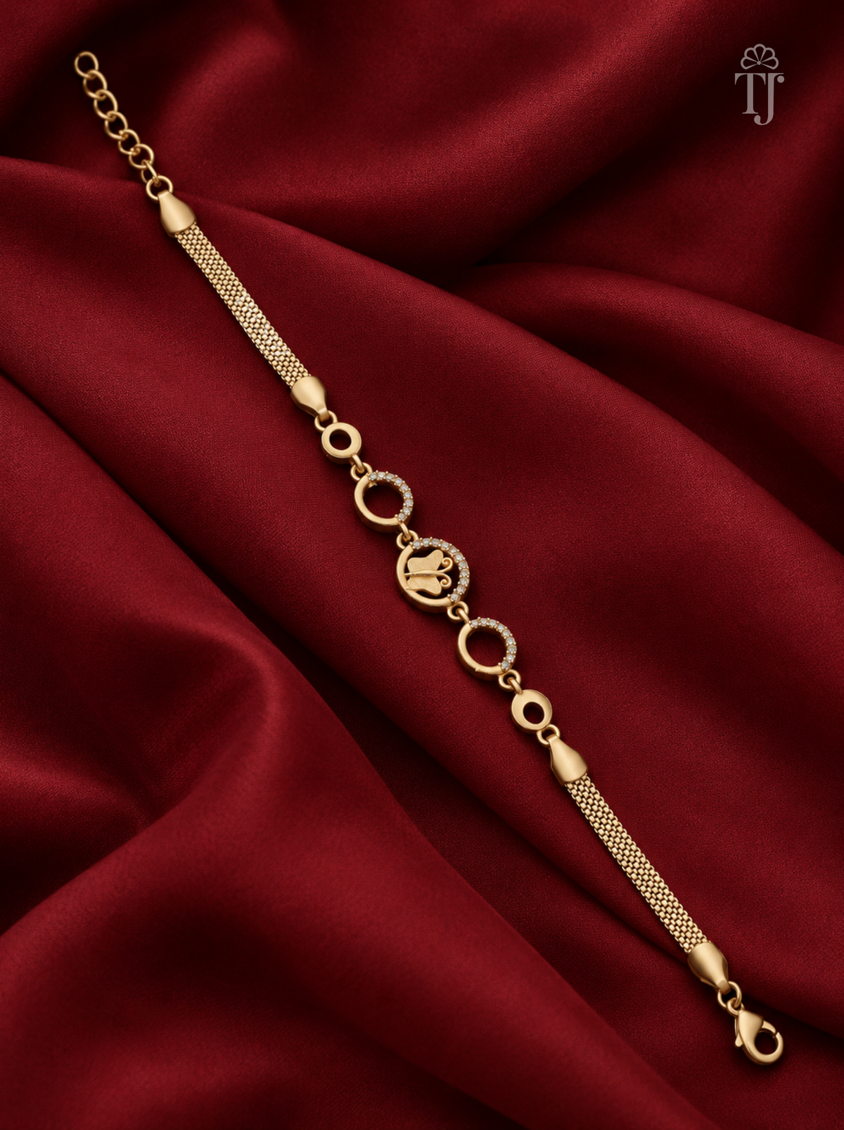 A gold-toned chain bracelet with a textured mesh section, circular links, and a central oval charm, displayed on draped deep red satin fabric.