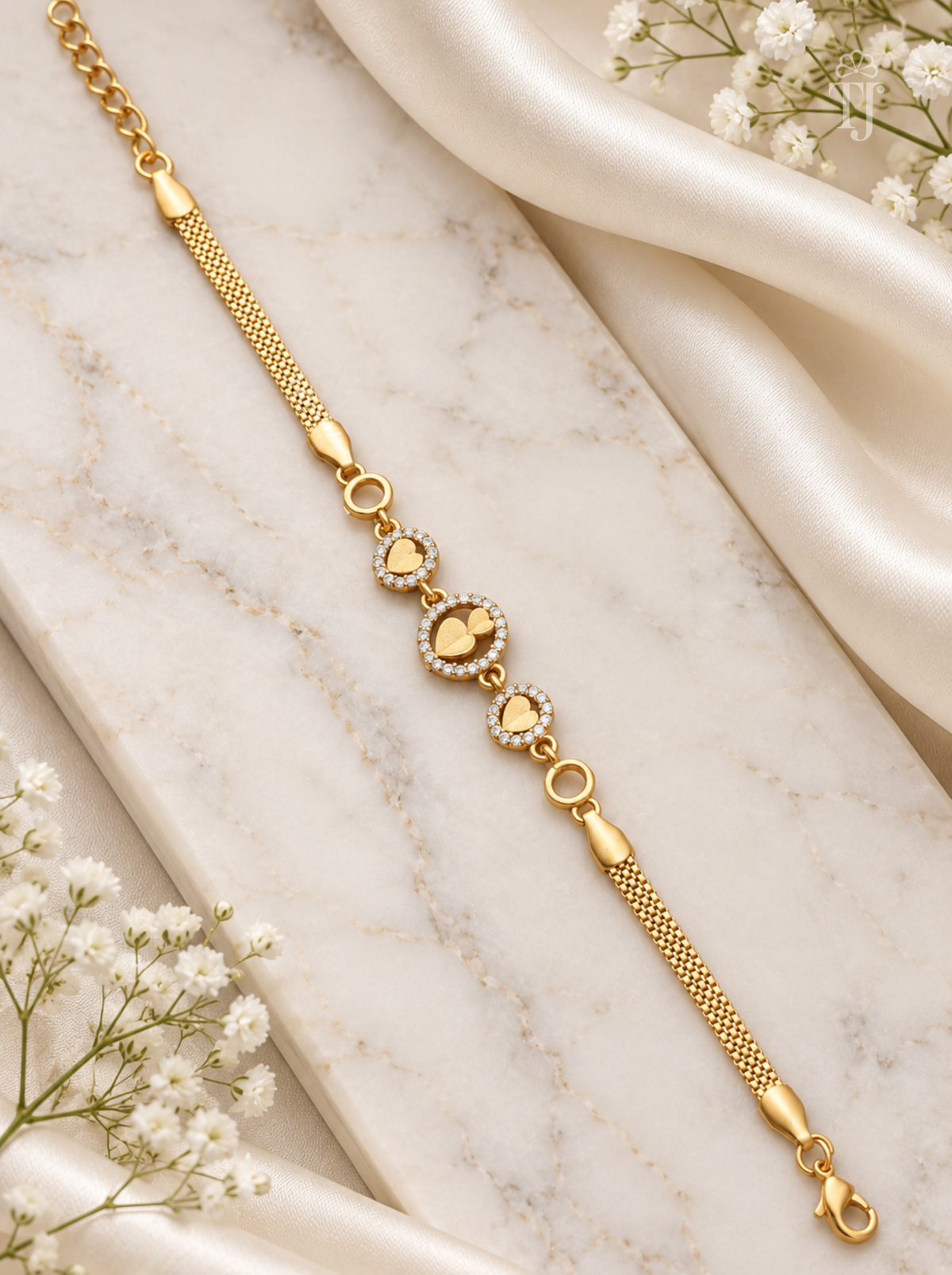 A gold chain bracelet with heart-shaped links, some adorned with small clear stones, displayed on a white marble slab with draped satin and white flowers.