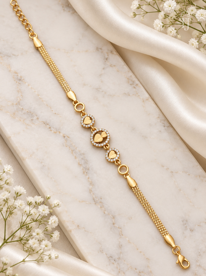 A gold chain bracelet with heart-shaped links, some adorned with small clear stones, displayed on a white marble slab with draped satin and white flowers.