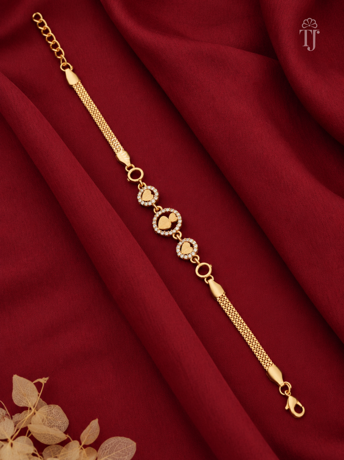 A gold chain bracelet with three heart-shaped motifs, some adorned with small clear stones, displayed on draped dark red fabric with dried leaves in the corner.