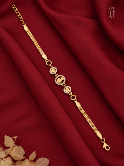 A gold chain bracelet with three heart-shaped motifs, some adorned with small clear stones, displayed on draped dark red fabric with dried leaves in the corner.