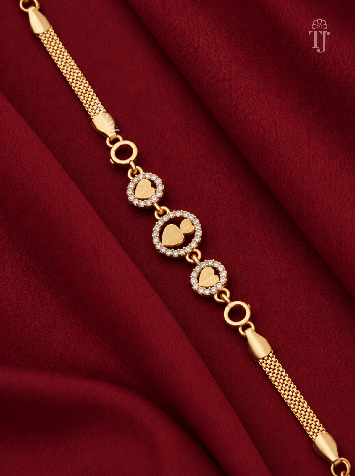 A gold chain bracelet with three heart-shaped charms, some adorned with small clear stones, displayed on a draped dark red fabric background.