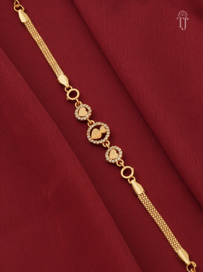 A gold-plated chain bracelet with three heart-shaped charms, some adorned with small clear stones, displayed on a draped dark red fabric background.