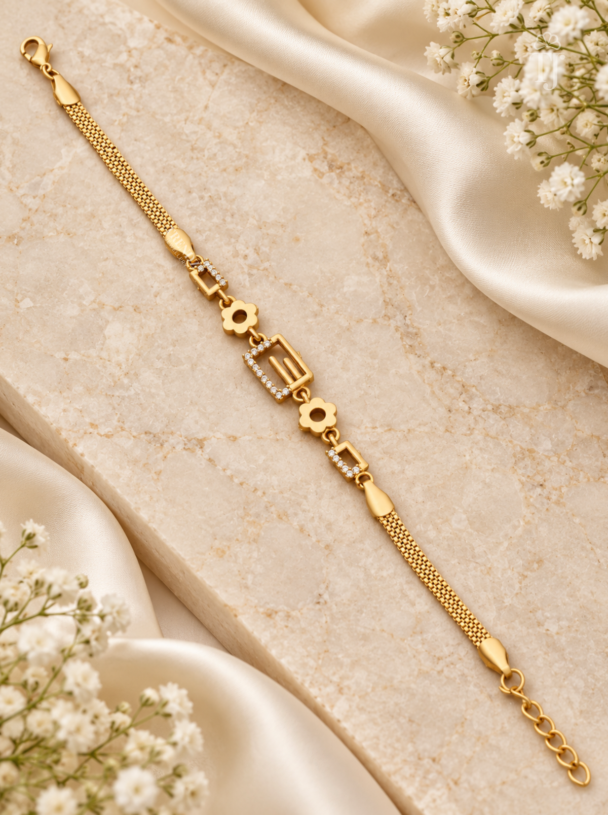 A gold chain bracelet with floral and geometric charms, styled on a beige stone slab with cream satin fabric and white baby's breath flowers.