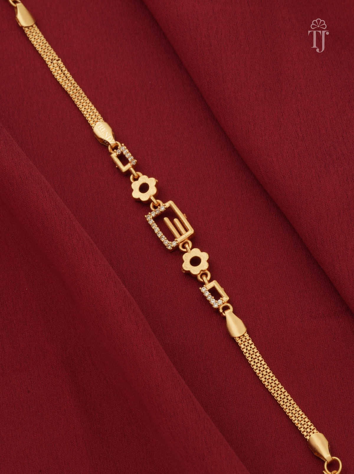 A gold-plated chain bracelet with geometric and floral motifs, some embellished with small clear stones, displayed diagonally on a draped dark red fabric background.