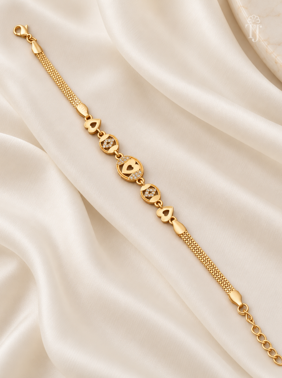 A gold chain bracelet with heart-shaped charms and clear gemstone accents, displayed on a draped cream satin fabric background.