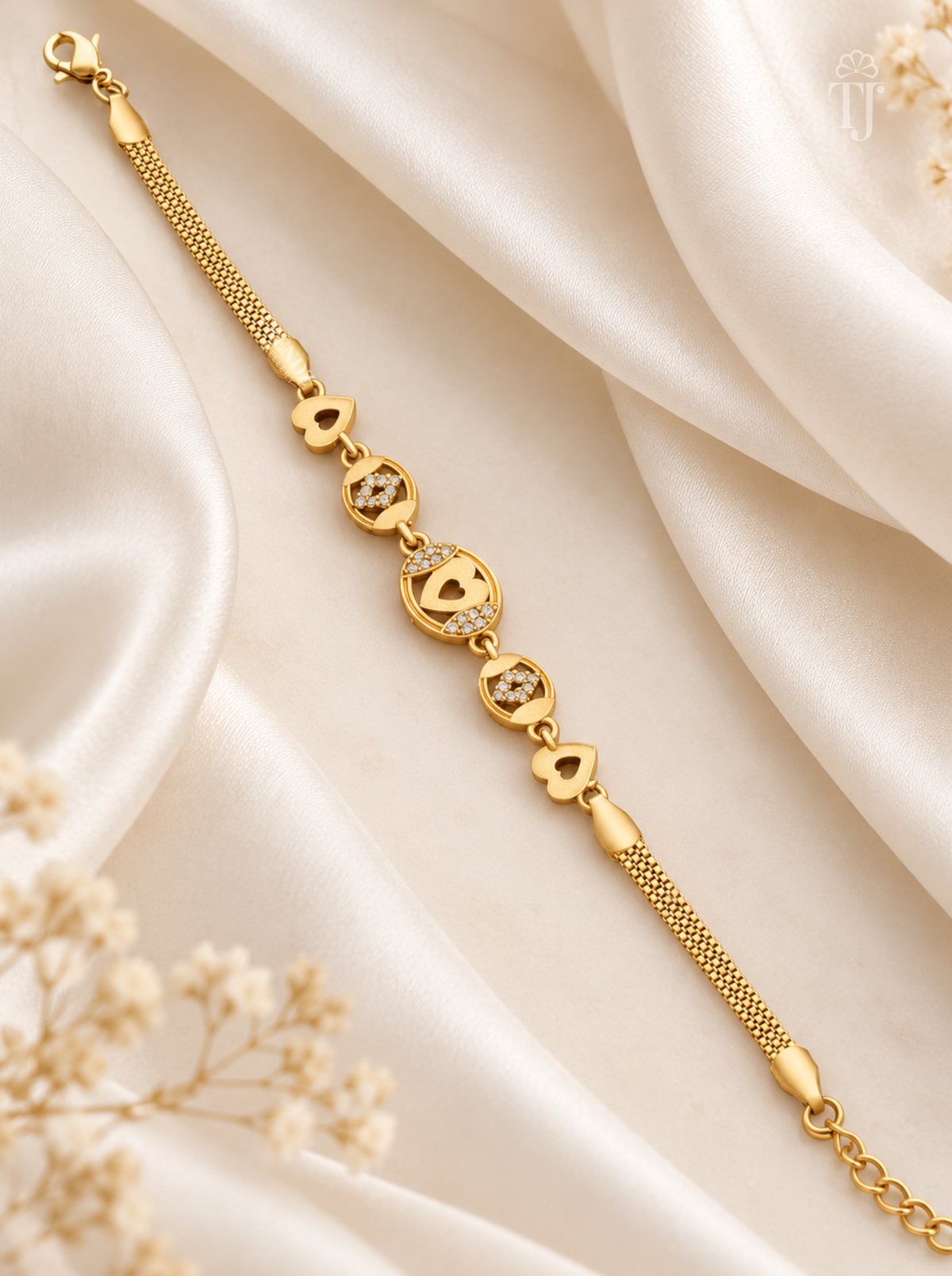 A gold chain bracelet with heart-shaped charms and small clear stones, displayed on draped cream satin fabric with dried floral accents.