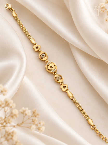A gold chain bracelet with heart-shaped charms and small clear stones, displayed on draped cream satin fabric with dried floral accents.