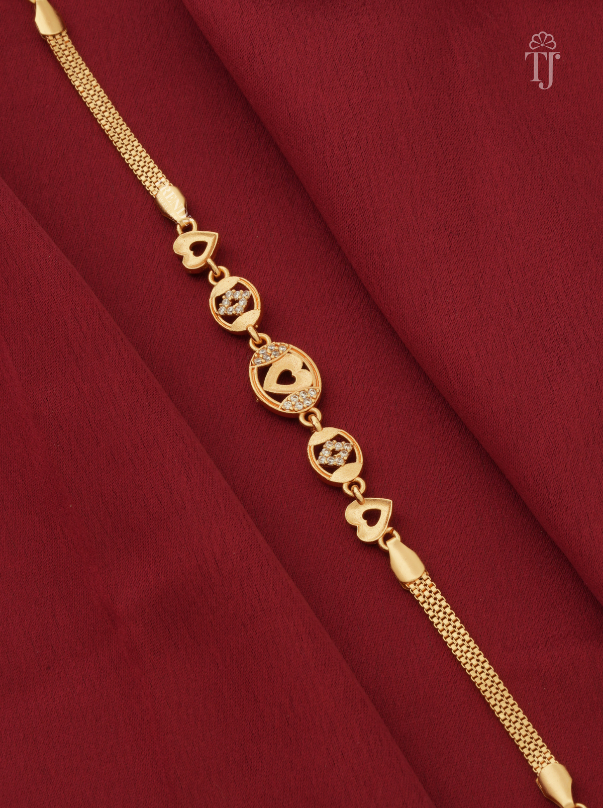 A gold-plated chain bracelet with heart and circular motifs, laid diagonally on a textured dark red fabric background.