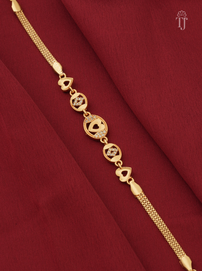 A gold-plated chain bracelet with heart and circular motifs, laid diagonally on a textured dark red fabric background.
